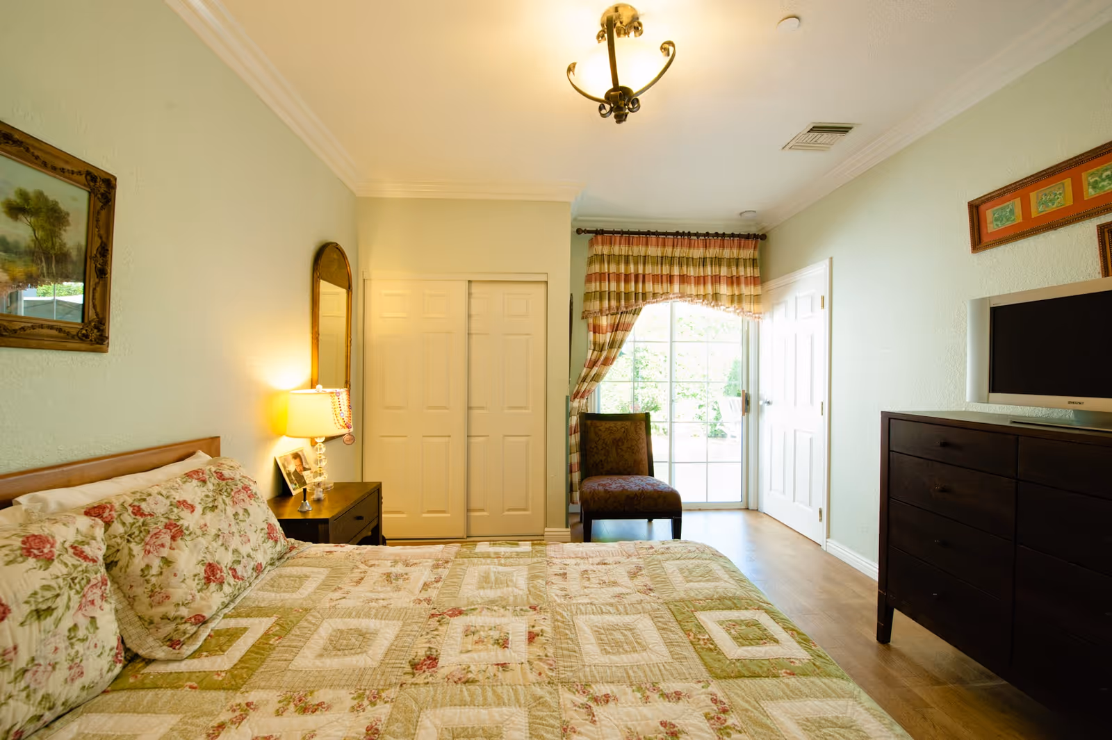 A cozy bedroom with a bed covered in a floral and patchwork quilt, matching pillows, a wooden nightstand with a lamp and framed photo, a large mirror, a chair near a sliding glass door with striped curtains, and a dark wooden dresser with a flat-screen TV on top.