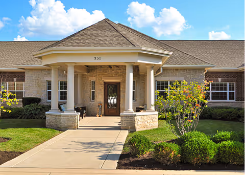 Front exterior view of a single-story building with a covered entrance supported by four columns. The building has a stone facade with a wooden door and windows on either side. There are neatly trimmed bushes and small trees along the walkway leading to the entrance under a blue sky with scattered clouds.