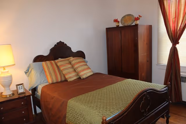 A cozy bedroom with a wooden bed featuring a carved headboard and footboard. The bed is made with brown and green bedding and striped pillows. To the left of the bed is a wooden nightstand with a white lamp and a small framed number 11. In the corner, there is a wooden wardrobe with decorative items on top. A window with red and orange striped curtains is on the right side of the room.
