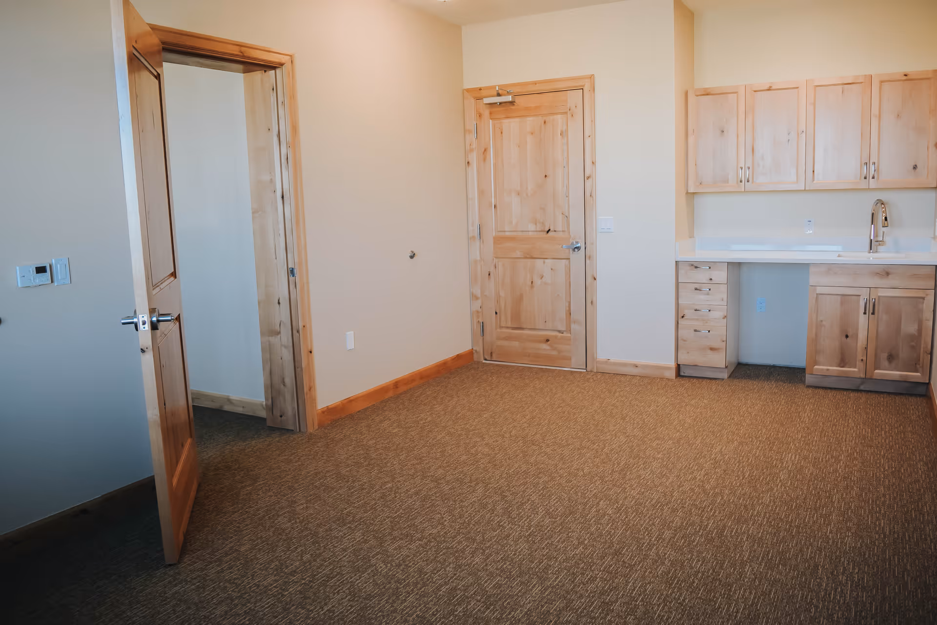 Empty room with beige walls and carpeted floor, featuring wooden doors and wooden cabinets with a white countertop and a sink on the right side.