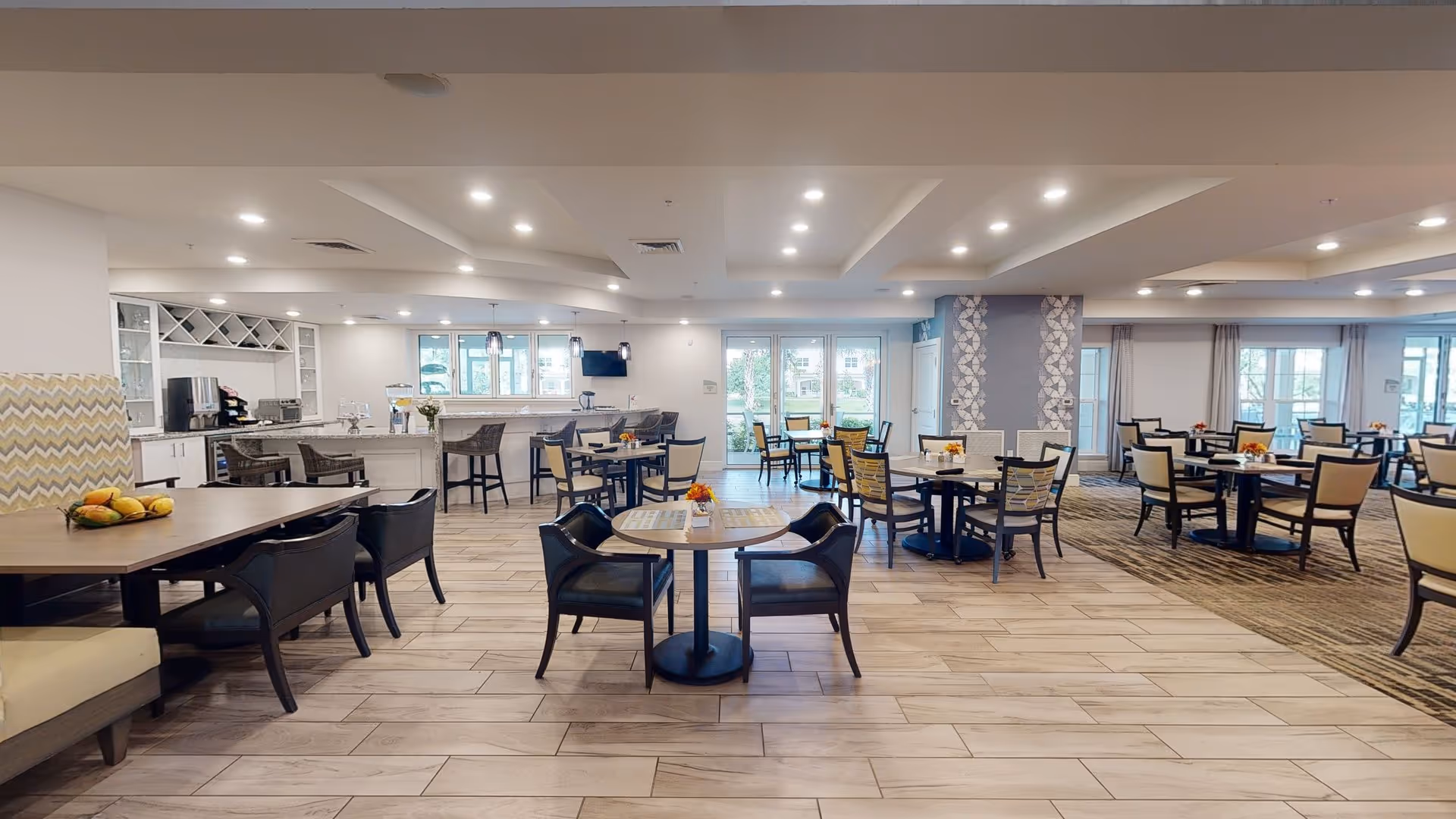 A spacious and well-lit dining area with multiple tables and chairs arranged neatly. The room features a combination of wooden and cushioned seating, a counter with bar stools, and large windows and glass doors allowing natural light to enter. The floor is tiled, and the ceiling has recessed lighting.