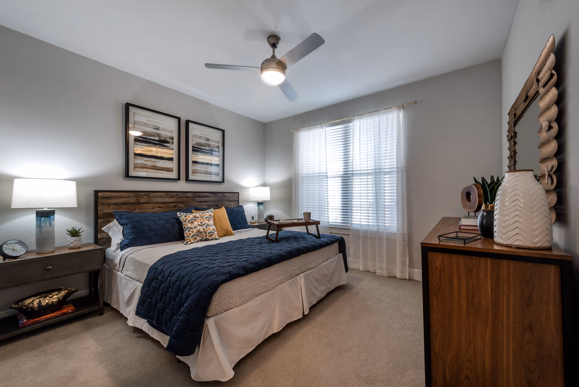 Well-lit furnished bedroom with a bed, nightstands, dresser, ceiling fan and a window with sheer curtains.
