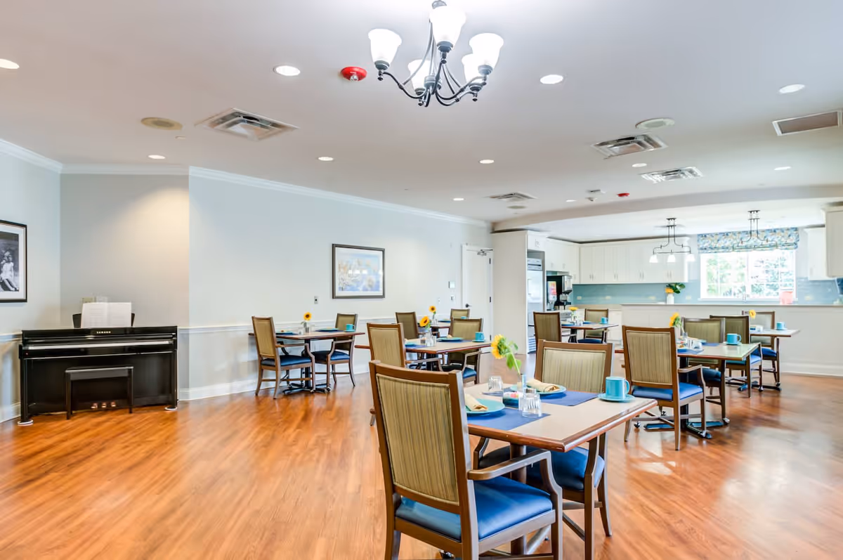 A bright dining room with several tables and chairs arranged neatly. Each table has place settings with plates, cups, and napkins, along with small vases holding sunflowers. The room features wooden flooring, light-colored walls, a piano against one wall, framed pictures, and a kitchen area with white cabinets and a window in the background. Ceiling lights and a chandelier provide illumination.
