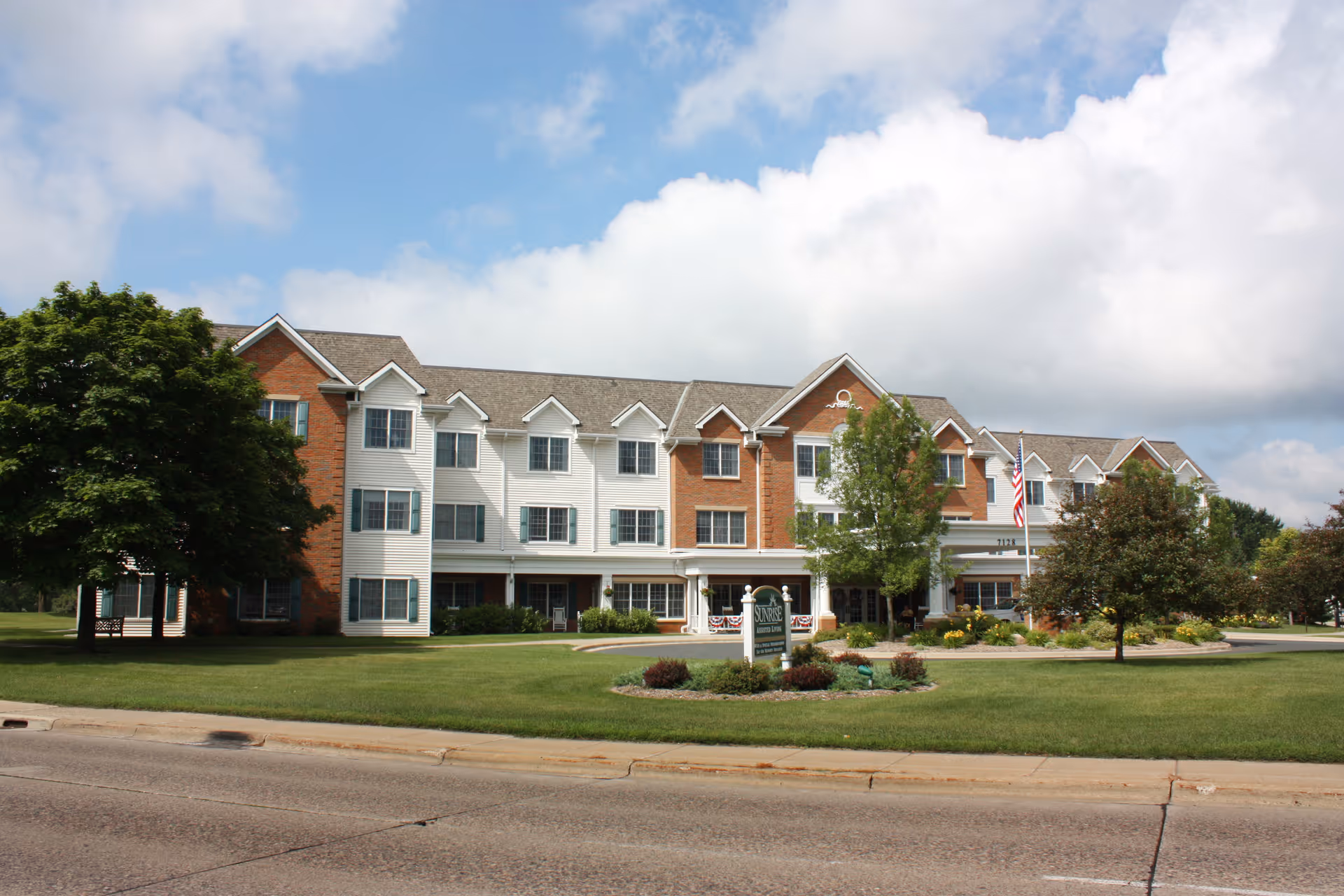 Exterior view of a three-story assisted living facility named Sunrise Of Edina with a mix of brick and white siding, surrounded by green lawns, trees, and a partly cloudy sky.