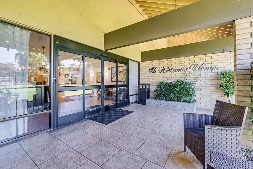 Covered entrance area of a senior living facility with sliding glass doors leading inside. There is a tiled floor, a black doormat, a brown wicker chair on the right, and a planter with green bushes against a brick wall with the words 'Welcome Home' written in cursive.