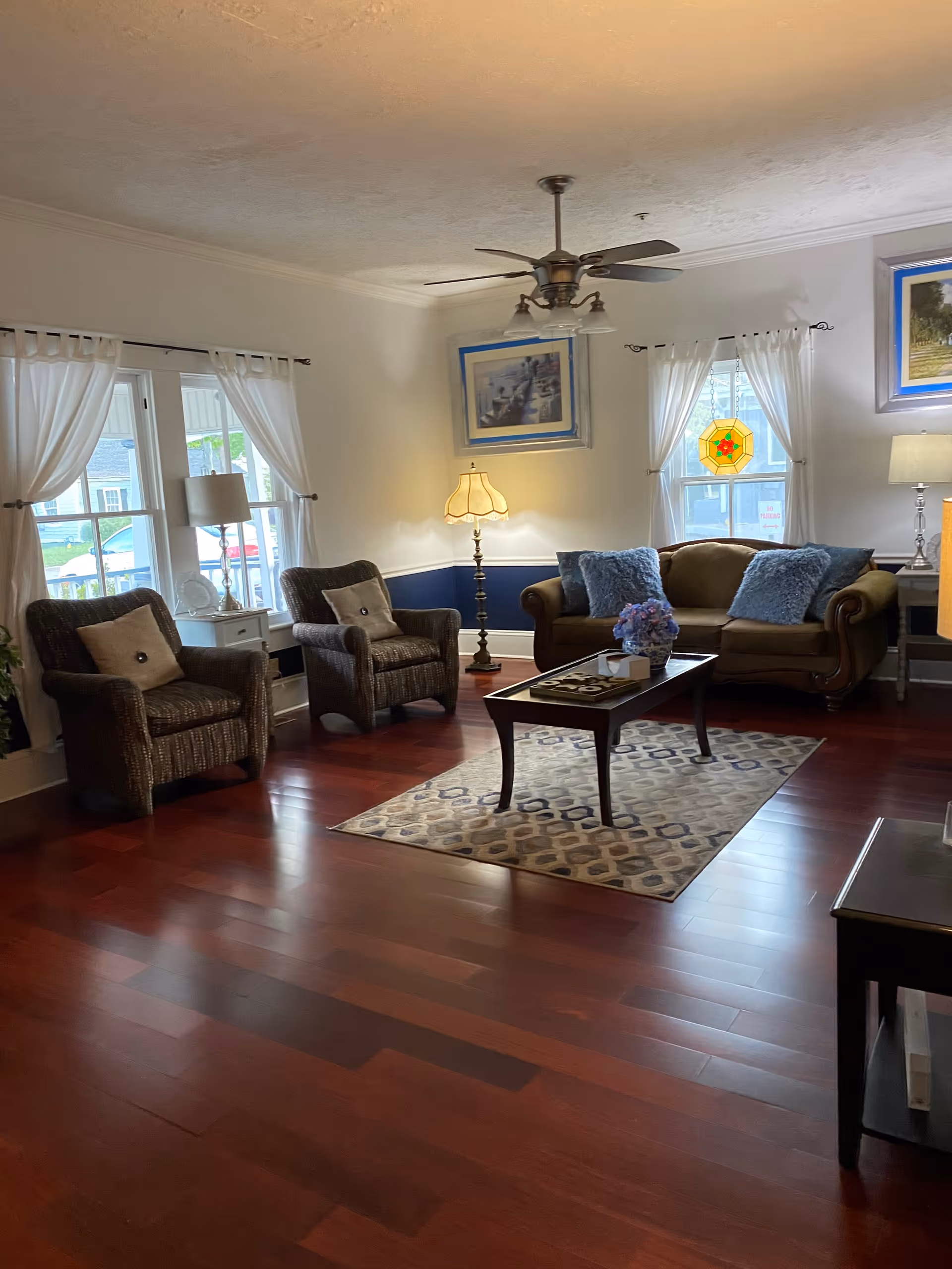 A cozy living room with polished wooden floors, a brown sofa with blue cushions, two wicker armchairs with beige pillows, a wooden coffee table with a floral centerpiece on a patterned rug, two windows with white curtains, a standing lamp, a ceiling fan with lights, and framed artwork on the walls.