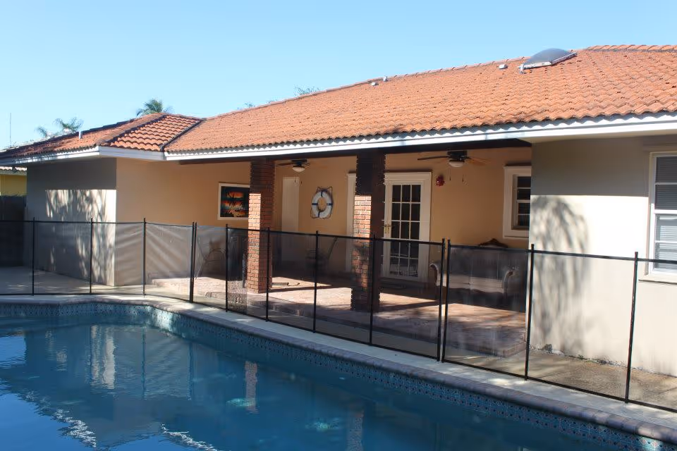 Outdoor view of a residential-style building with a red tiled roof and beige walls, featuring a covered patio area with brick pillars and ceiling fans. In front of the patio is a swimming pool enclosed by a black safety fence.