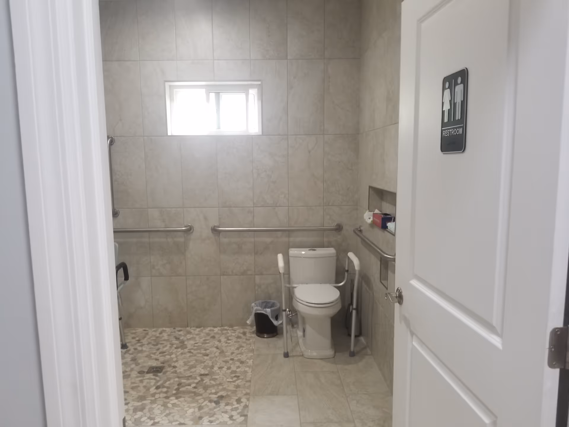 View into a restroom with beige tiled walls and floor. The restroom features a toilet with safety rails on both sides, a small window above, a trash can, and a recessed shelf holding tissue boxes. There are grab bars mounted on the walls for accessibility. The door has a restroom sign showing male and female icons.