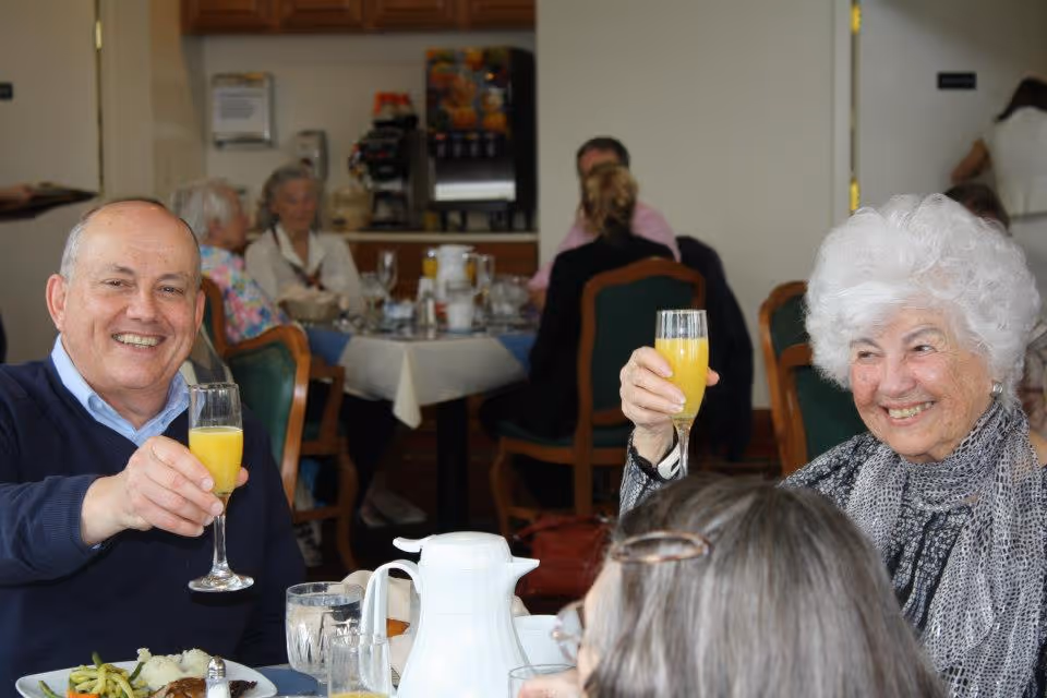 Two smiling older adults at a dining table raising glasses of orange beverage in a communal dining room.