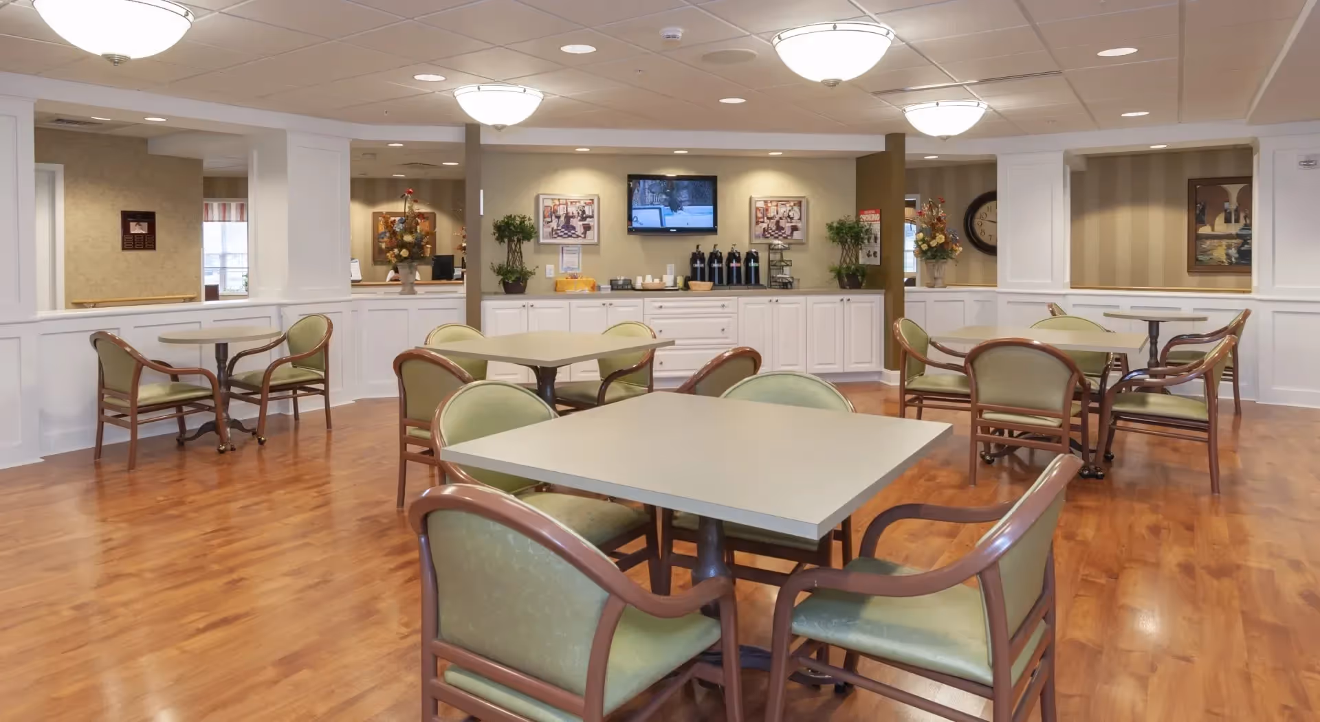 A spacious dining area in an assisted living facility with several square tables surrounded by green cushioned chairs. The room has wood flooring, white wainscoting on the walls, and soft overhead lighting. At the back, there is a counter with coffee dispensers, cups, and a wall-mounted TV above it. Decorative plants and framed pictures adorn the walls.