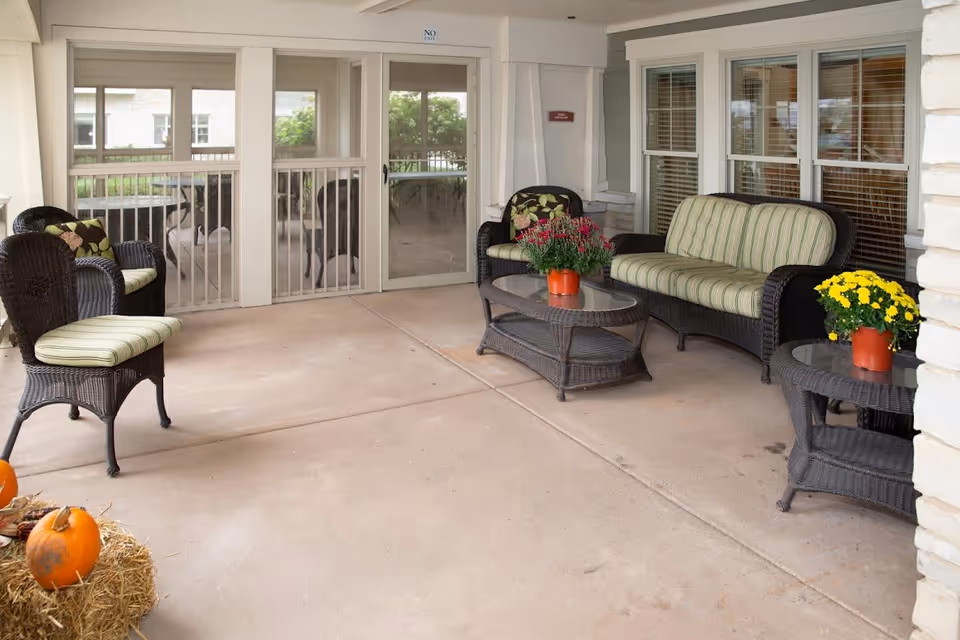 Covered outdoor patio area with wicker furniture including a cushioned loveseat, chairs, and glass-top tables. There are potted flowers on the tables and pumpkins on a bale of hay in the corner. Sliding glass doors and windows with blinds are visible in the background.