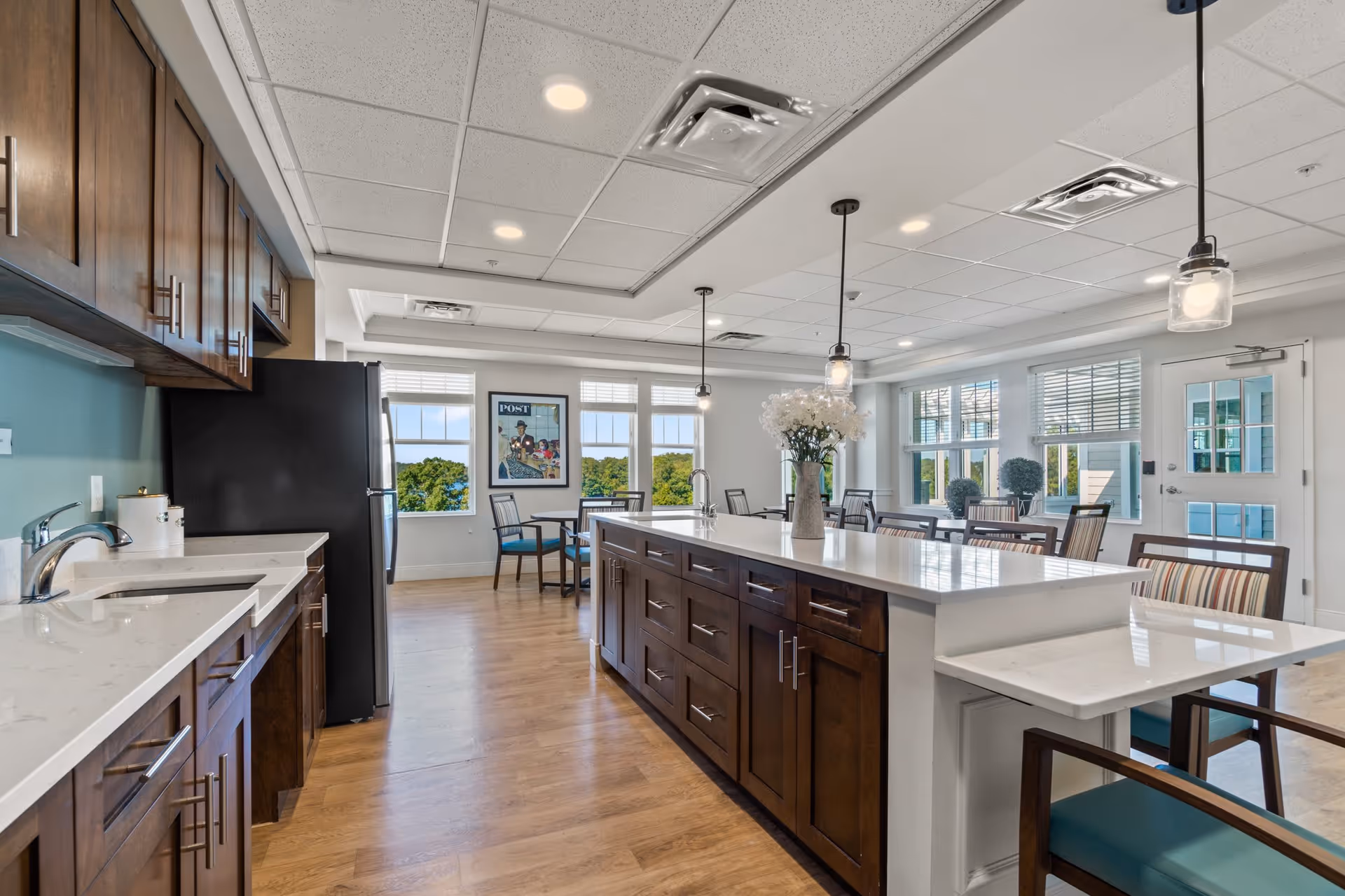 Bright and spacious kitchen and dining area in a senior living facility featuring wooden cabinets, a large central island with a white countertop, pendant lights hanging from the ceiling, and multiple tables and chairs near large windows letting in natural light.