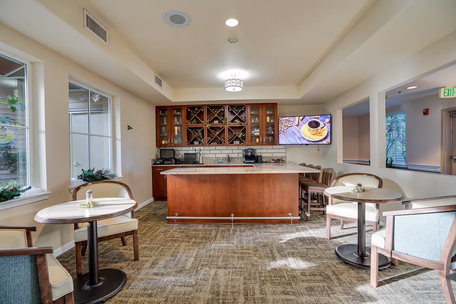 A cozy common area with a central wooden island countertop and wine rack cabinetry behind it. The room has carpeted flooring, two small round tables with chairs on either side, large windows letting in natural light, and a wall-mounted TV displaying a coffee cup image. The space is well-lit with ceiling lights and decorated with small plants.