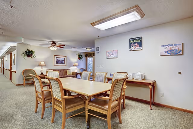 A well-lit communal dining area with a rectangular wooden table surrounded by chairs, a sofa and wall decorations.