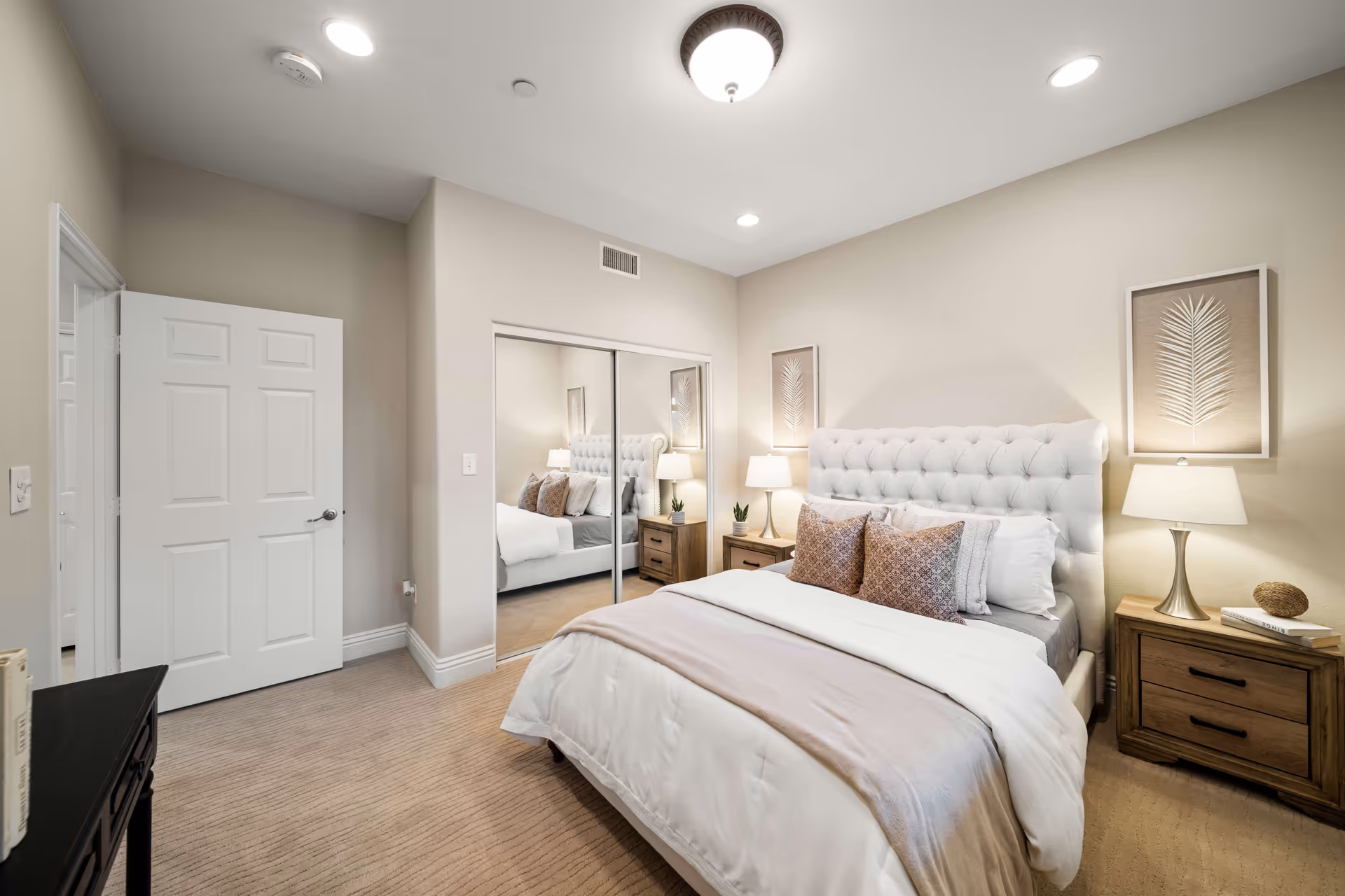 A neatly made bed with a tufted white headboard in a bedroom. The bed has white and beige bedding with two decorative pillows. On each side of the bed are wooden nightstands with lamps and small decorative items. There is a large mirrored closet door on the wall opposite the bed, and a white door is open to the left. The walls are painted beige and the carpet is light brown.