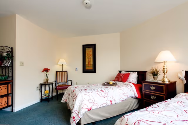 A bright bedroom with two twin beds featuring white bedspreads with red floral patterns. Between the beds is a wooden nightstand with a brass lamp and a small plant. On the left side of the room, there is a small round table with a vase of red flowers and a striped chair. A framed floral artwork hangs on the wall above the bed. The room has light-colored walls and a green carpeted floor.