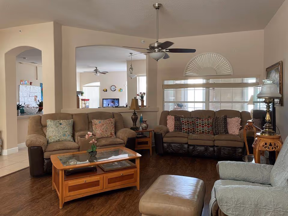 A cozy living room with two brown sofas adorned with colorful pillows, a glass-top wooden coffee table, a beige armchair with a matching ottoman, and a wooden side table with a vintage lamp. The room has wooden flooring, beige walls, a ceiling fan, and large windows with blinds allowing natural light to enter.