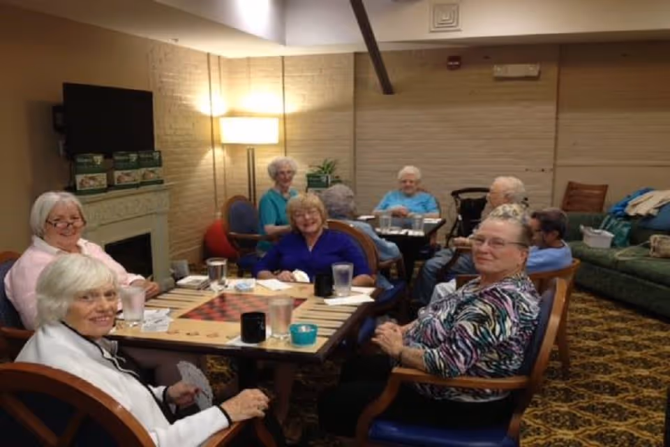 A group of elderly people sitting around tables in a cozy room playing card games and socializing. The room has warm lighting, a patterned carpet, a green couch, and a television mounted on the wall above a fireplace.
