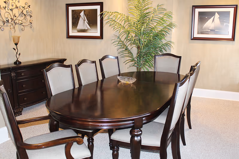 Elegant dining room with a dark wood oval table surrounded by upholstered chairs, framed sailboat pictures, a sideboard and a potted plant.