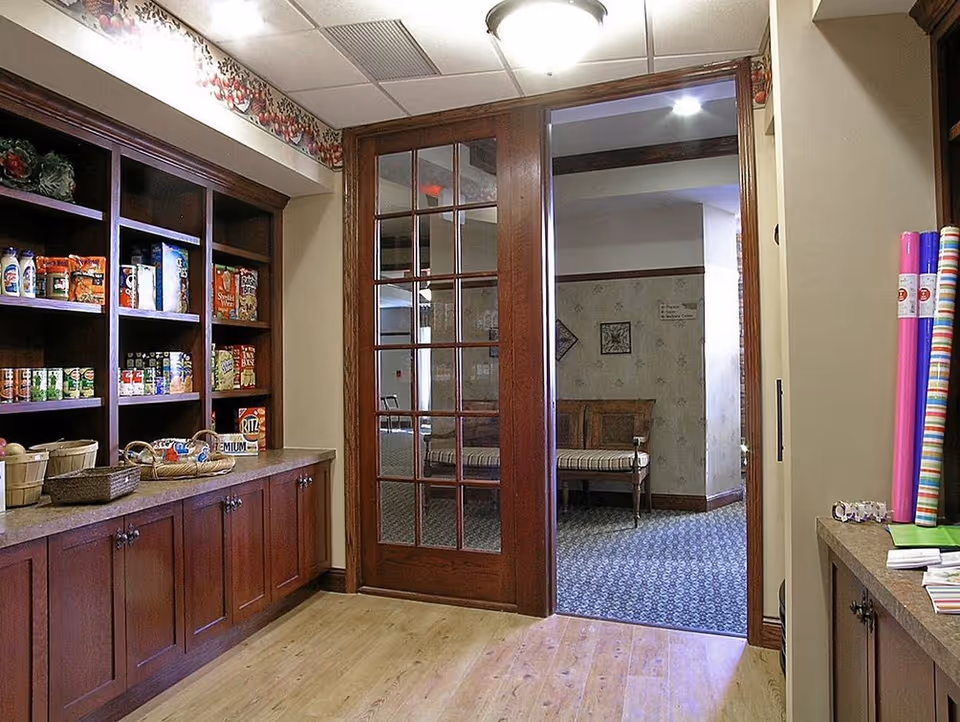 A pantry area with wooden shelves stocked with various canned and boxed food items on the left side, and a countertop with baskets and other items. There is a wooden framed glass door leading to a carpeted room with a wooden bench and framed wall decorations.