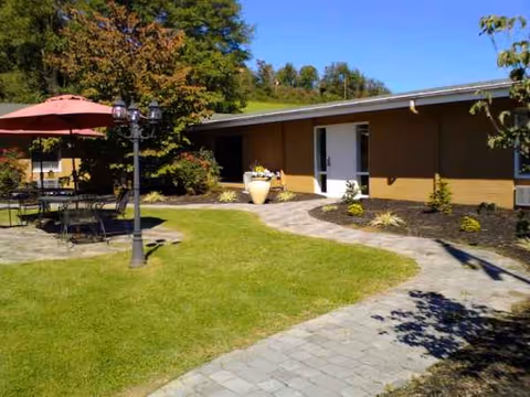 Outdoor courtyard with a single-story brown building, green lawn, and patio seating under a red umbrella.