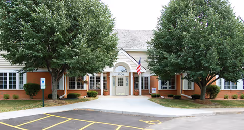 Front exterior view of Brookdale Middleton Stonefield facility showing a single-story building with a central entrance, two large trees flanking the walkway, an American flag near the door, and a parking area with handicap parking spaces in the foreground.
