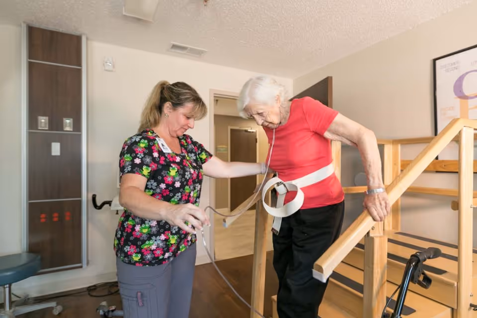 A healthcare worker assists an elderly woman wearing a red shirt and black pants as she uses wooden handrails to practice walking. The elderly woman is wearing a medical belt and has a nasal cannula for oxygen support. They are in a rehabilitation center room with medical equipment and a poster on the wall.