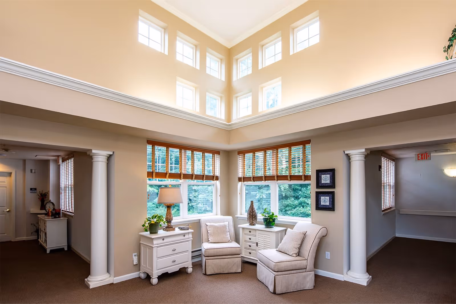 A bright and cozy sitting area in a senior living facility with two cushioned chairs, a small white side table with a lamp and plants, large windows with wooden blinds, and high windows near the ceiling allowing natural light to fill the space. The area is framed by two white columns and beige walls.