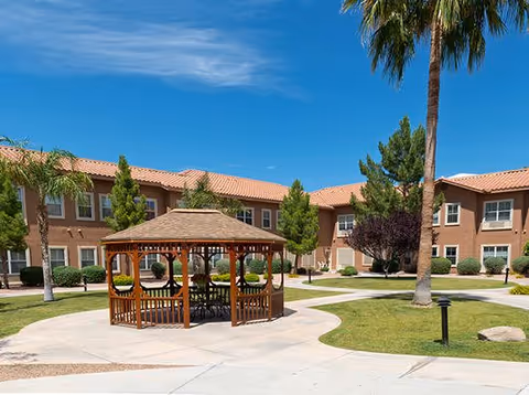 Sunlit courtyard with a wooden gazebo, palm trees, manicured lawn, and a two-story building in the background.