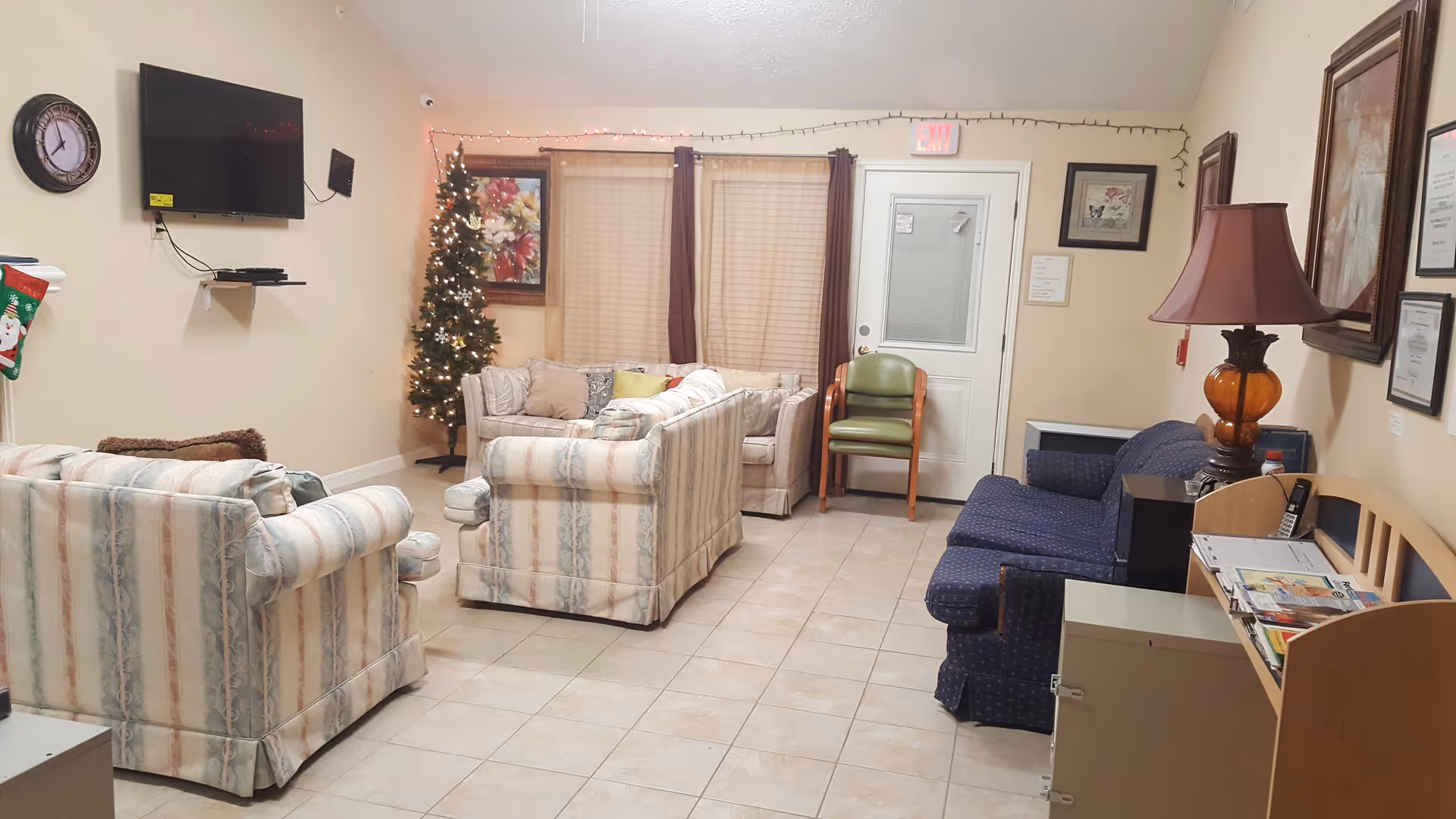 A cozy living room with tiled floor, featuring two patterned sofas, a blue couch, a green chair, a small decorated Christmas tree, a wall-mounted TV, a clock, framed pictures, and a lamp on a side table. There is a door with a window and curtains on the windows.
