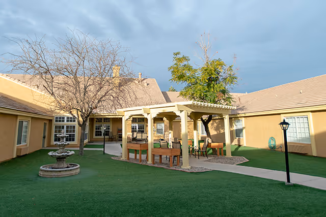 Outdoor courtyard area at Brookdale North Gilbert featuring a green lawn, a multi-tiered water fountain, a covered pergola with seating, leafless and leafy trees, and surrounding beige buildings under a cloudy sky.