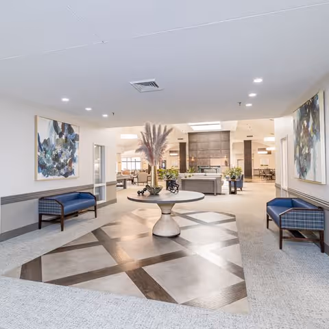 A spacious and well-lit common area in a senior living facility featuring a round table with decorative plants in the center. The room has two blue cushioned benches with plaid upholstery on either side, abstract paintings on the walls, and a fireplace in the background. The flooring includes a patterned section in the middle surrounded by carpet.