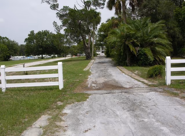 A cracked concrete driveway lined by white split-rail fencing, grass and palm trees leading toward a building in the distance.