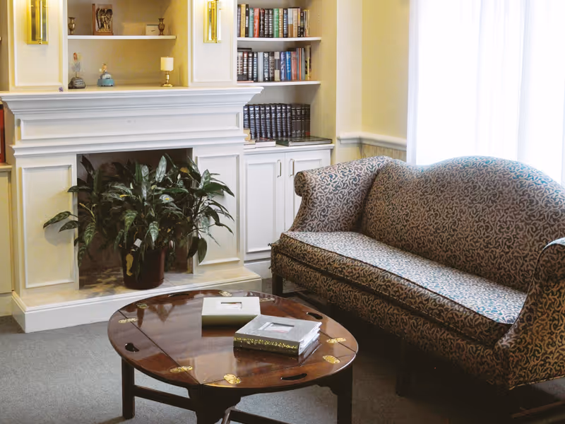 A cozy sitting area with a patterned sofa, wooden coffee table holding books, and a white fireplace with a potted plant and built-in bookshelves.