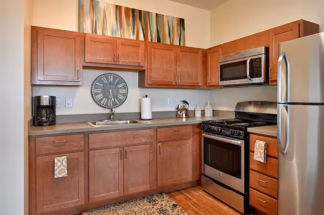 Compact kitchen with wooden cabinets, stainless steel refrigerator and stove, sink, coffee maker, and a large wall clock beneath decorative artwork.