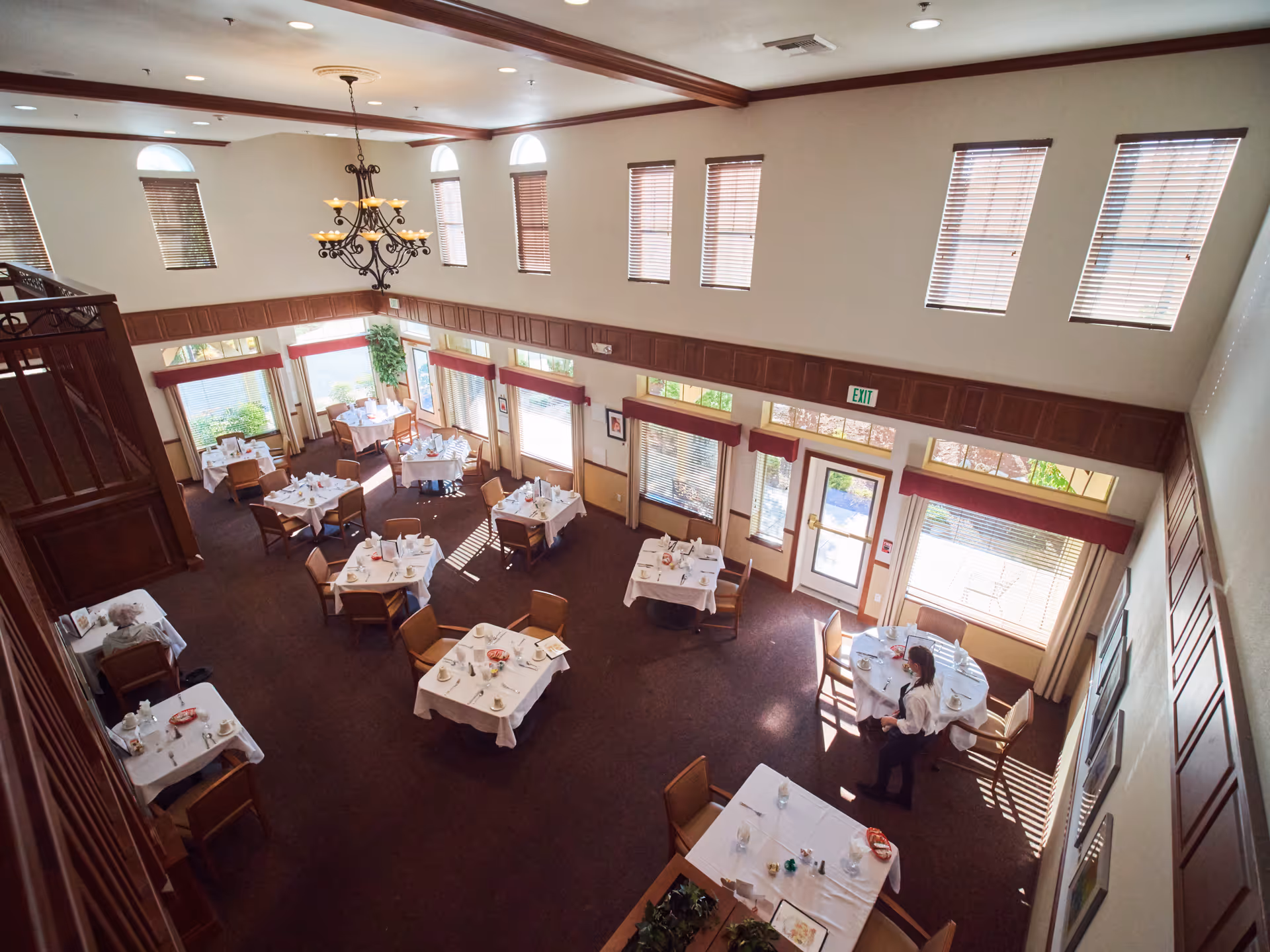 A spacious dining room with multiple tables covered in white tablecloths, set with plates, cups, and utensils. There are several chairs around each table, and a few people are seated or standing. The room has high ceilings with several windows letting in natural light, wooden paneling along the walls, and a chandelier hanging from the ceiling.