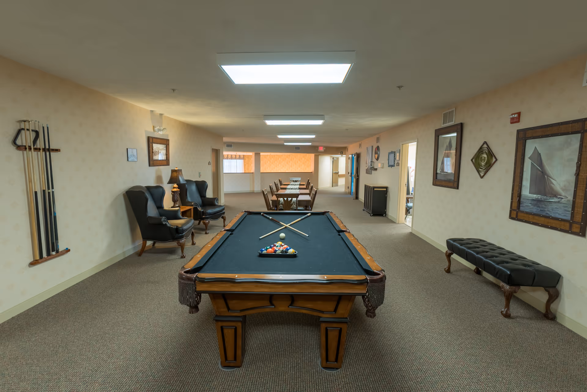 Interior view of a recreational room in a senior living facility featuring a pool table with cues and balls arranged, two black armchairs with a small table and lamp between them on the left, a long table with chairs in the background, and framed artwork on the walls.
