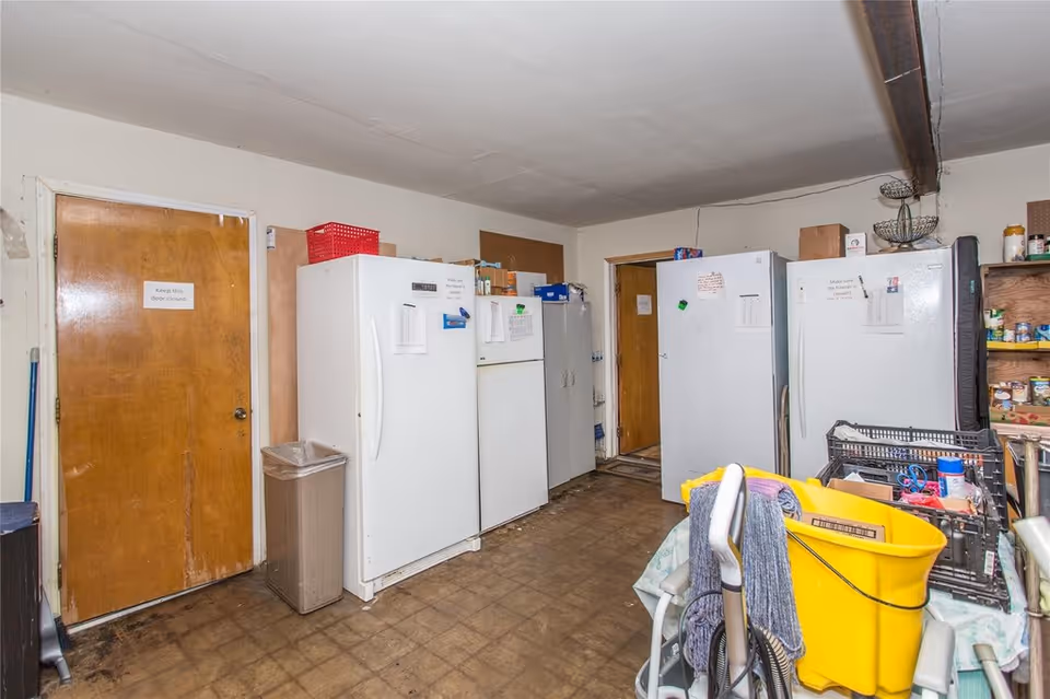 Utility/storage room with several upright freezers and refrigerators, shelving of canned goods, and cleaning supplies including a yellow mop bucket.