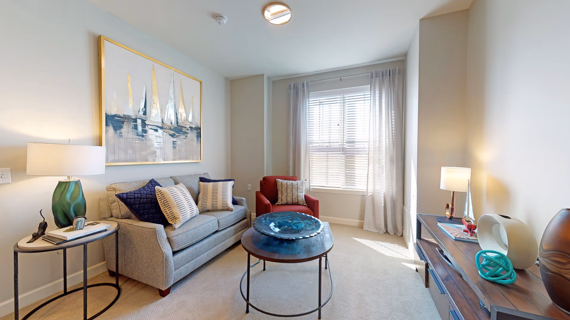 A bright living room with a gray sofa adorned with blue and white pillows, a red armchair, a round wooden coffee table with a decorative blue bowl, a side table with a green lamp and small decor items, a wooden console table with a lamp and decorative objects, and a window with white blinds and sheer curtains allowing natural light to fill the room. A large painting of sailboats hangs above the sofa.