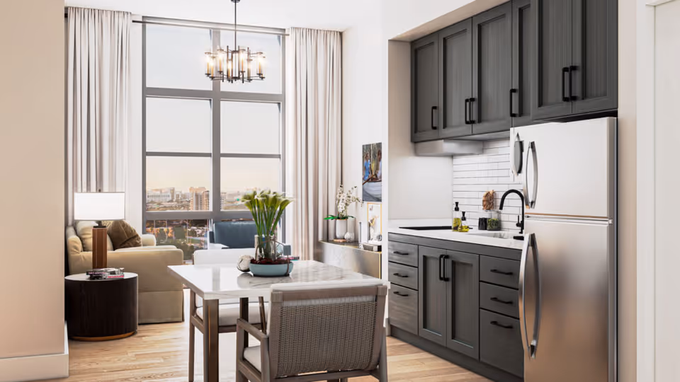 Bright open-plan kitchen and dining area with a marble table and chairs, dark gray cabinets and a stainless refrigerator beside a living area by a large window.