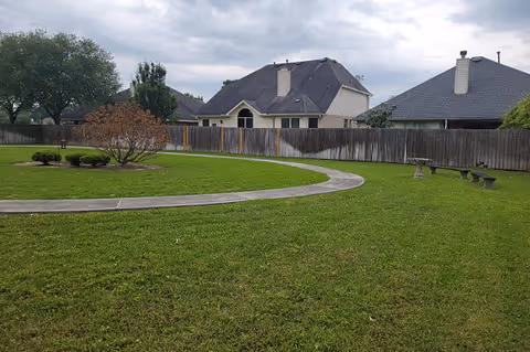 A grassy outdoor area with a curved concrete walkway, a few small bushes and trees, a wooden fence, and houses in the background under a cloudy sky.