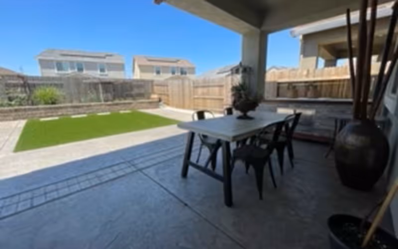 Covered backyard patio with a dining table and chairs, potted plants, and a fenced yard with artificial turf under a clear sky.