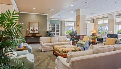 A spacious and well-lit living room area in a senior living facility with multiple beige and green upholstered sofas and armchairs arranged around a large floral-patterned ottoman. The room features large windows with curtains, several potted plants, a wooden console table with decorative items and a mirror above it, and built-in shelves with books and decor.