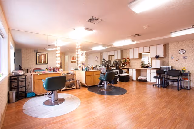 Interior view of a salon area in a senior living facility with two salon chairs placed on circular mats in front of large mirrors with lights. The room has wooden flooring, cabinets along the back wall, and various salon equipment and supplies on countertops and shelves.