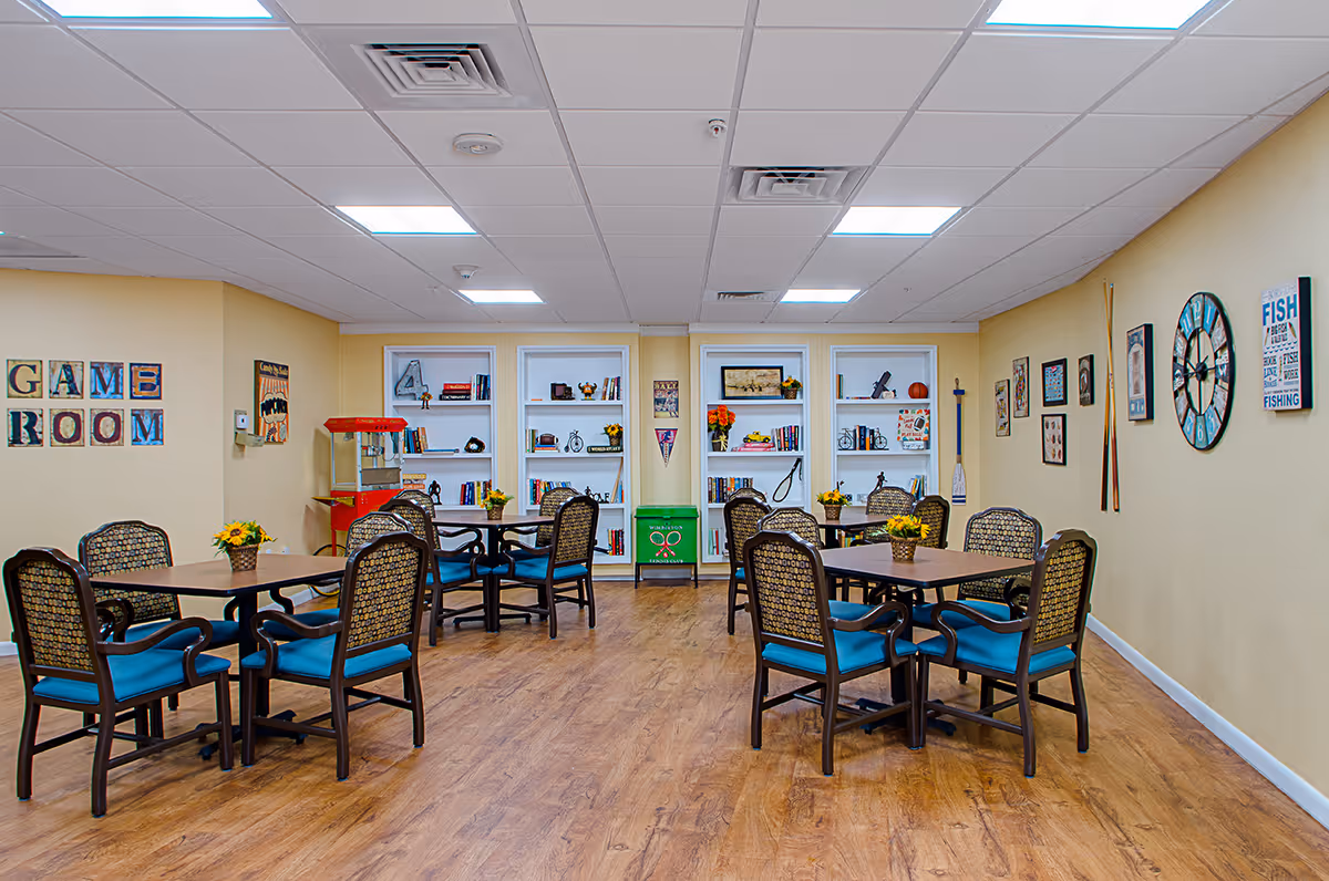 Spacious game room with several tables and blue-upholstered chairs, bookshelves, wall decor and a popcorn machine.