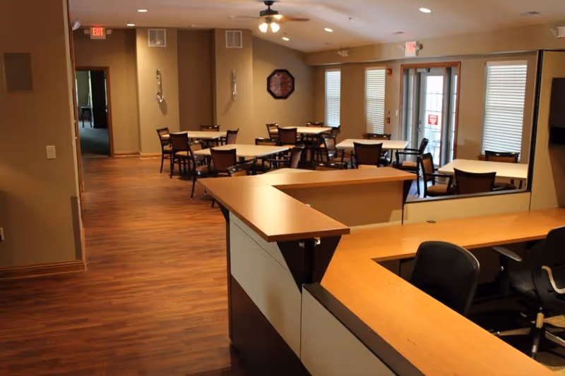 Interior view of a senior living facility dining area with multiple tables and chairs arranged neatly. The room has wooden flooring, beige walls, and several windows with blinds. A reception desk with office chairs is visible in the foreground, and ceiling fans with lights are mounted on the ceiling.