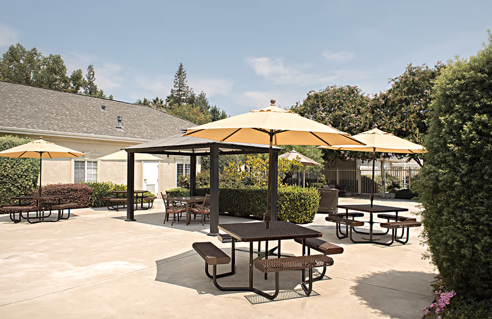 Outdoor patio area with multiple picnic tables and benches, each shaded by large beige umbrellas. The area is surrounded by greenery including bushes and trees, with a building visible in the background under a clear sky.
