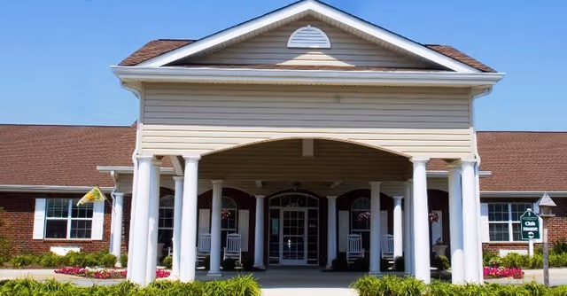 Front exterior view of a single-story building with a covered entrance supported by white columns, a brown shingled roof, and a clear blue sky. There are flower beds and greenery in front of the building, and a sign indicating 'Club House' on the right side.