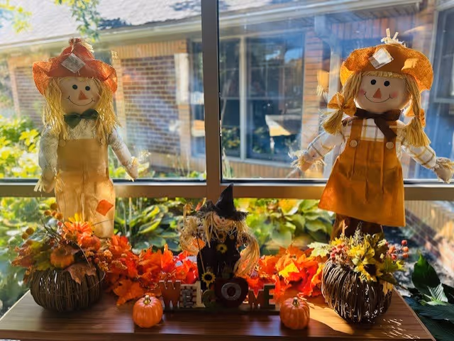 A window sill decorated with two scarecrow dolls dressed in autumn-themed clothing, surrounded by artificial fall leaves, small pumpkins, and a wooden 'WELCOME' sign with a small scarecrow figure in the center. The background shows a brick building and greenery outside the window.