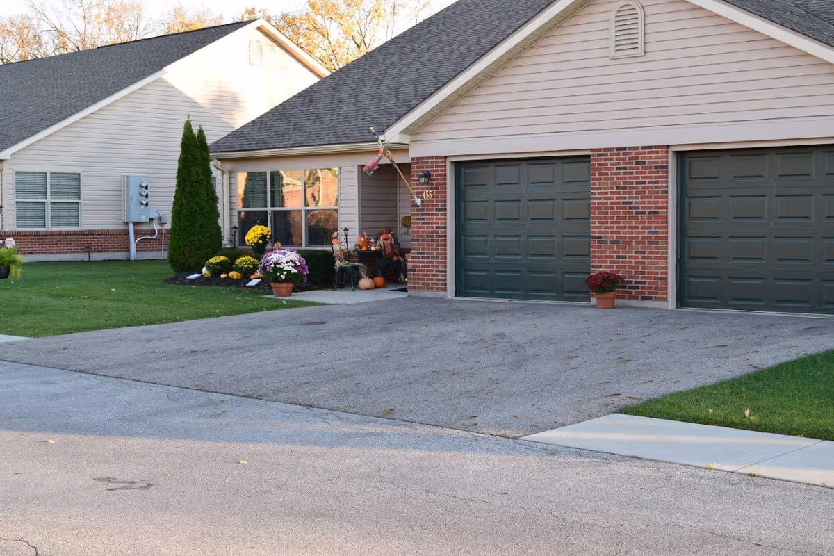 Exterior view of a residential building with two green garage doors, a driveway, and a small porch decorated with potted flowers and autumn-themed decorations including pumpkins. The building has beige siding and red brick accents.